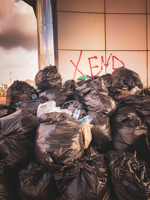 A large pile of black rubbish bags filled with waste, stacked against a tiled building wall outdoors. Some bags are torn open, revealing plastic bottles, cardboard, and assorted waste materials inside