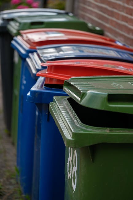 A red metal skip positioned against a dark green tiled wall and a light grey concrete wall, with the rear door of the skip closed. Several white plastic rubbish bags filled with waste are piled on the