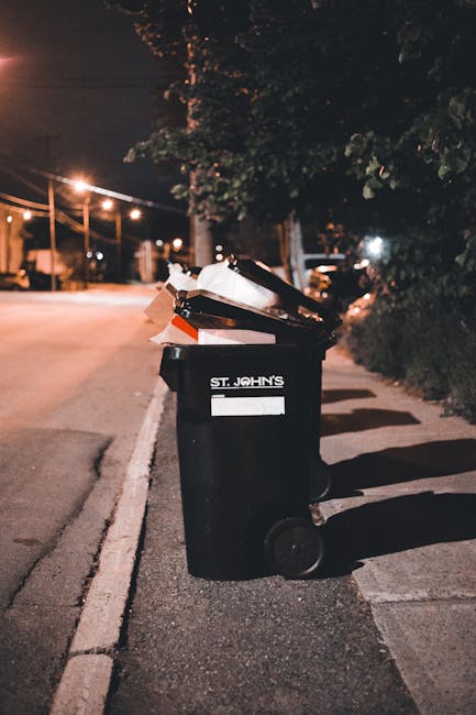 A man wearing a navy blue T-shirt with printed white text is leaning forward while placing a large white plastic bag filled with rubbish into a cylindrical metal recycling bin outdoors. The bin, made